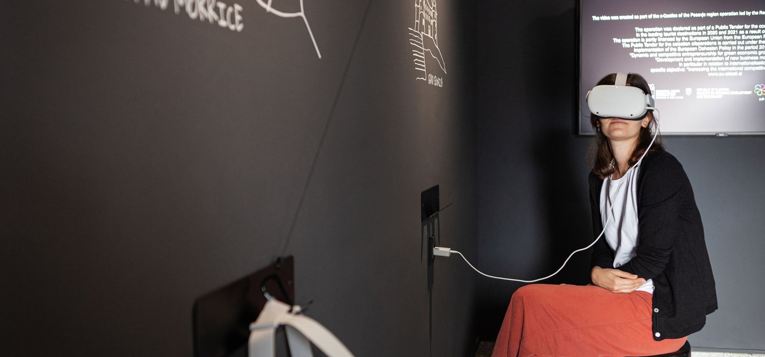 A woman sitting in a dark room wearing VR goggles and viewing a castle in the digital room e-Castles of Posavje.