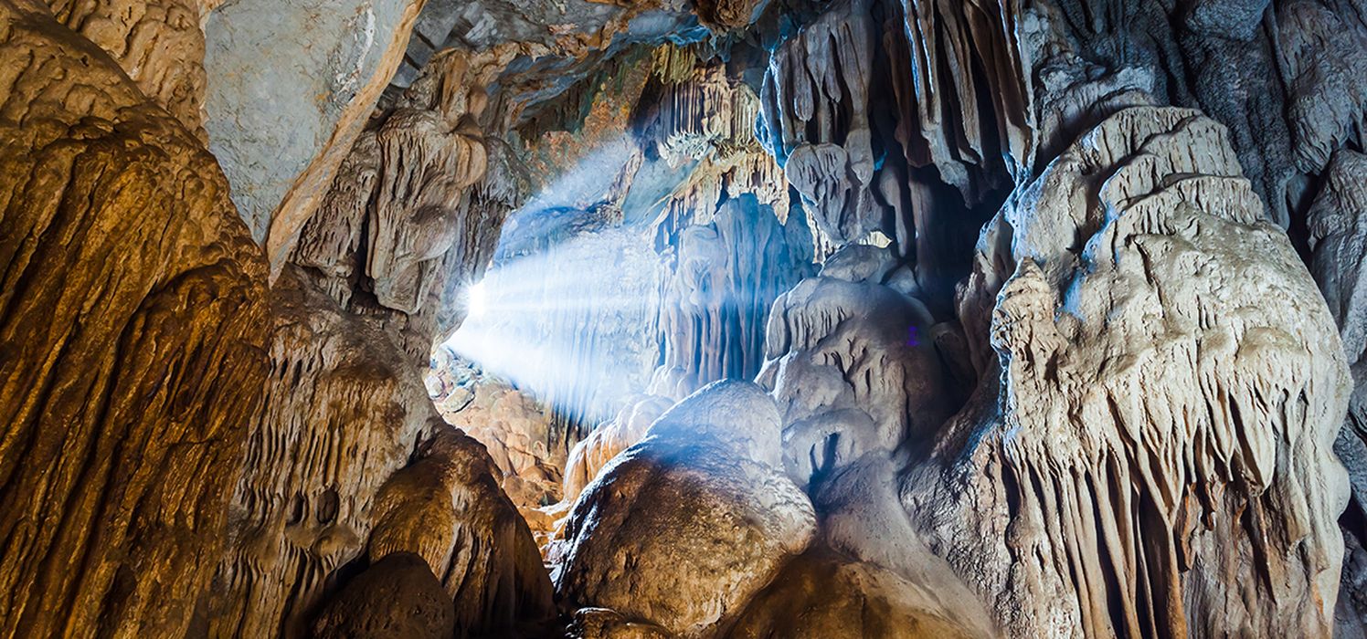 A photograph taken in Postojna Cave shows sunlight streaming through a hole in the wall, surrounded by stalactites.