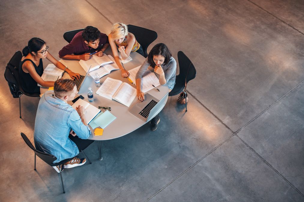 Top down photo of a group of people sitting around a white circular table. They have books, notebooks and a laptop.