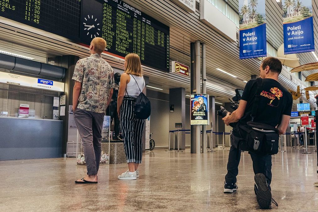 Photograph of an Arctur employee taking a photo of a two tourists looking at the arrival and departure board at the airport.