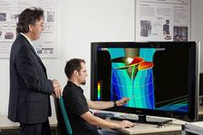 Photograph of a man sitting behind the computer showing a simulation of a turbine to the man standing behind him and listening.