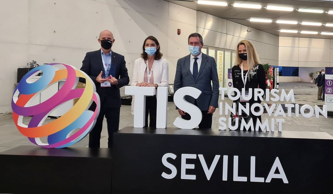 A group photograph of four people at the Tourism Innovation Summit. All the participants are wearing protective masks and are standing in front of the TIS logo.
