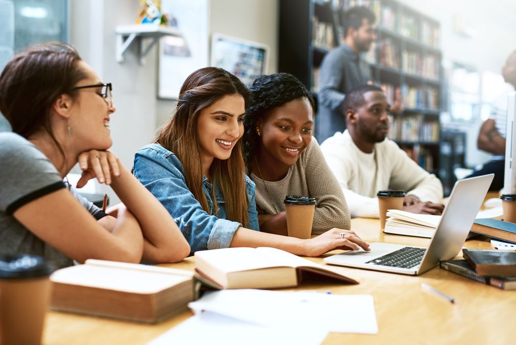 Photo of people in a library. In the front are three women looking at a laptop, smiling.