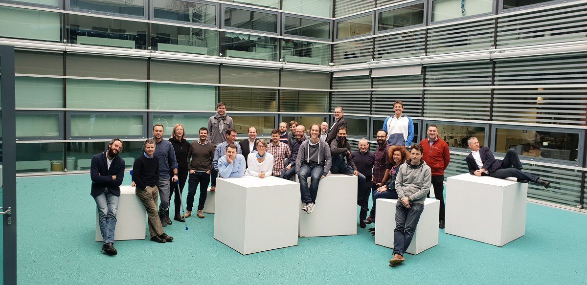 A group photograph of twenty-five people. They are standing and sitting on the white cubes various sizes. Behind them is a lass office building.