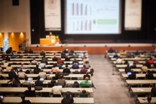 Photograph of a lecture hall where a group of listeners are attentively listening to a presenter. In the background is a large presenting screen with a presentation.