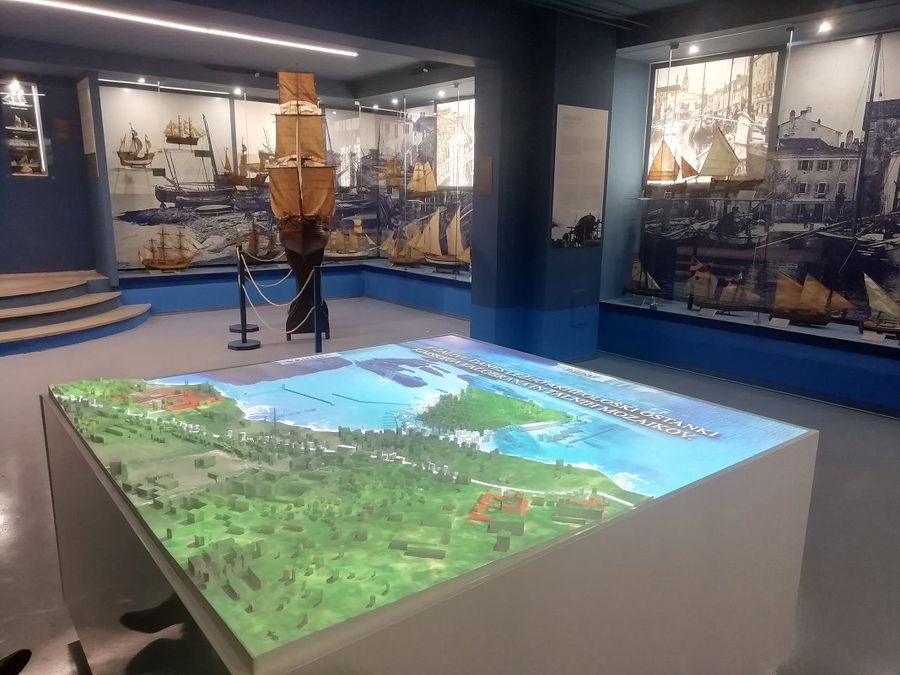 Photograph of a museum room where sail boats are displayed. In the middle of the room there is a geographical model of a town, forest and ocean.