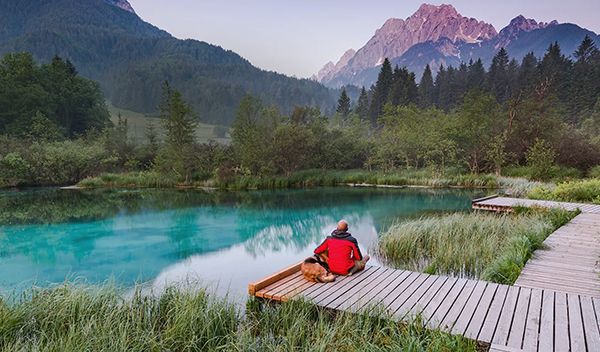 Photograph of a man and a dog sitting on a pier next to a lake, staring at the sunset-lit mountains in the background.
