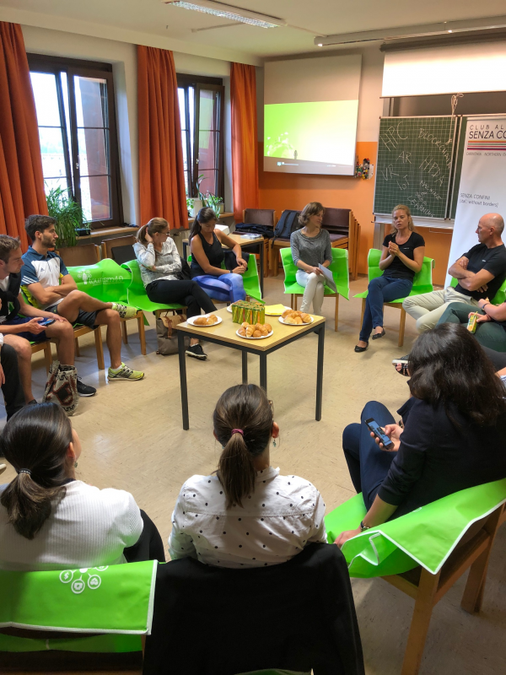A roundtable conversation. In the middle of of the circle of people is a table with croissants and orange juice.  