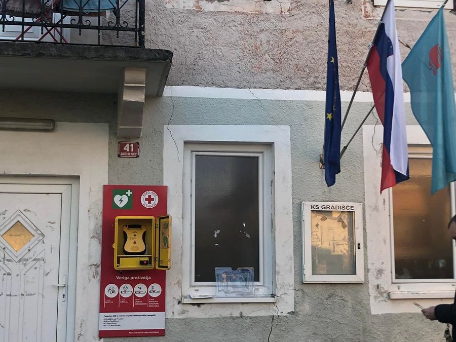 A photograph of an old house with a defibrillator hanging on it. There are also three flags hanging from the wall: the flag of the EU, the flag of Slovenia, and the flag of the municipality of Nova Gorica.