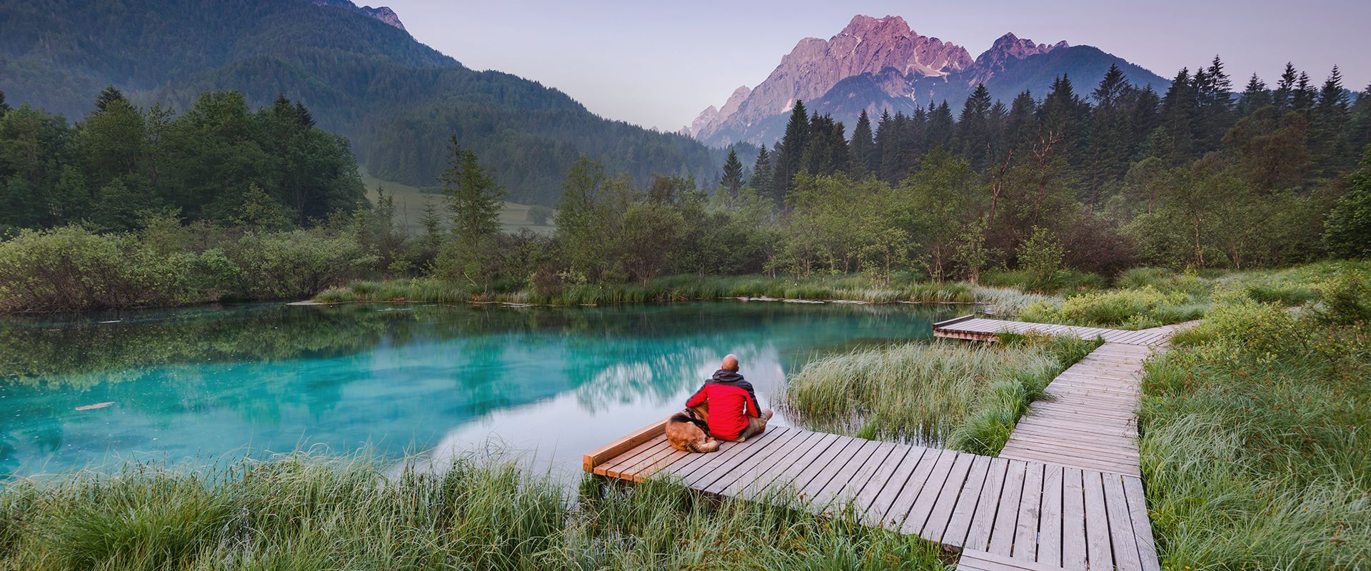 A man and a dog sitting on the pier near a lake, looking at the mountains.